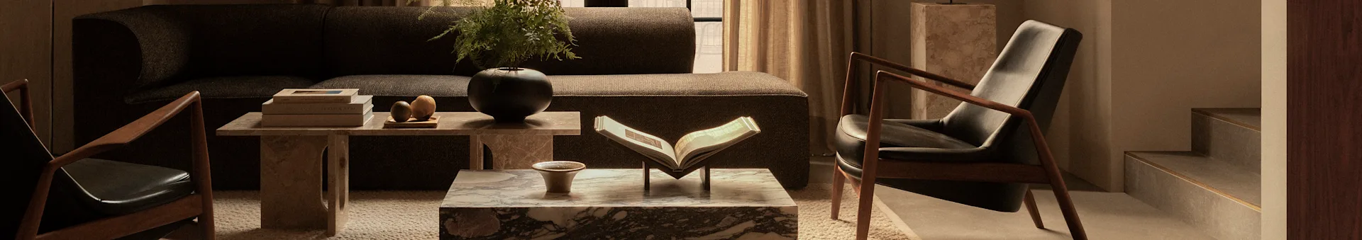 Een gezellige woonkamer met een donkerbruine bank, twee donkere leren fauteuils met houten frames, en marmeren salontafels versierd met boeken en planten.