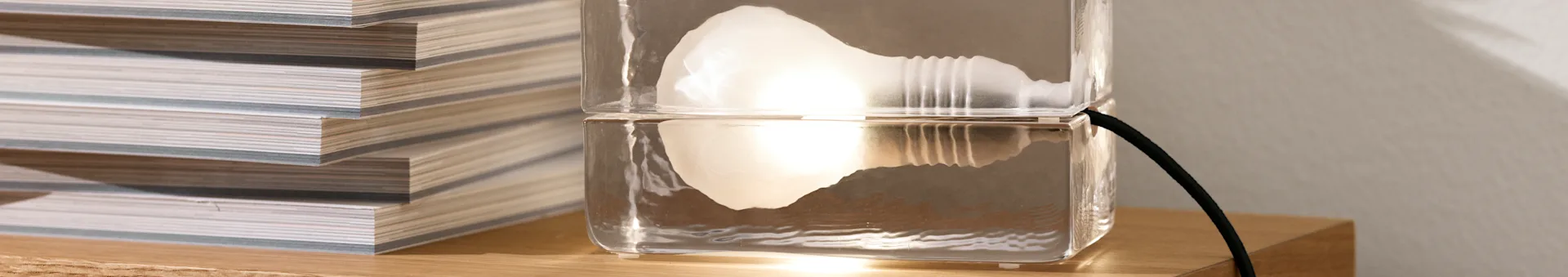 Een unieke heldere lamp, gemaakt van twee gestapelde rechthoekige blokken, met een gloeiend intern licht, naast een stapel tijdschriften op een houten oppervlak.