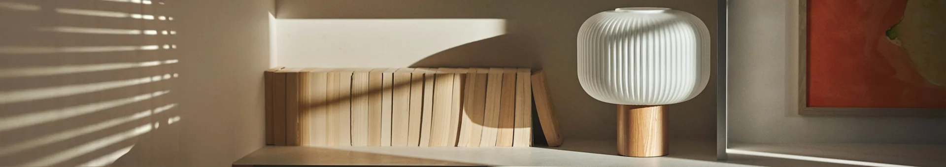Een moderne witte geribbelde lamp met een houten voet staat naast een rij lichte houten blokken, met zonlicht dat horizontale schaduwen werpt.