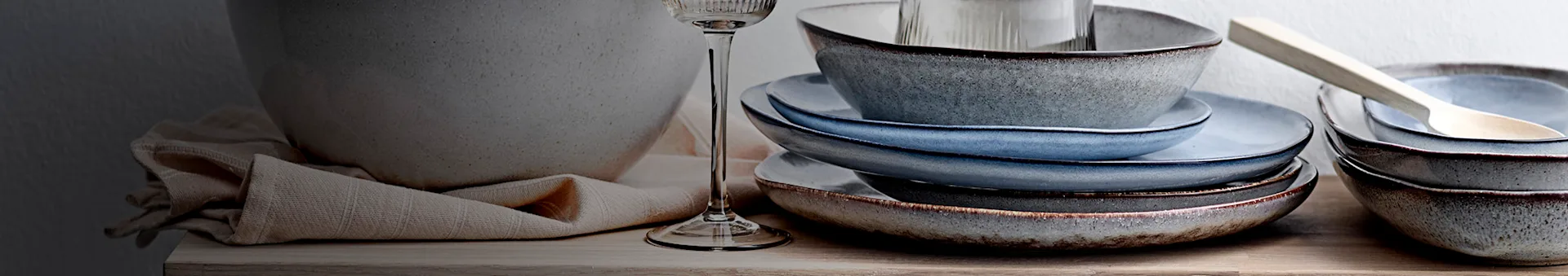 Gestapelde rustieke keramische kommen en borden in blauw- en grijstinten op een houten ondergrond, met glas en servet.