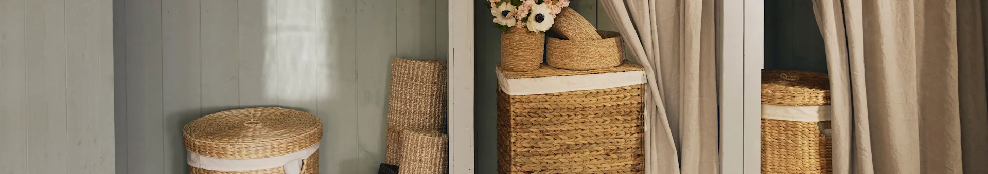 Natuurlijk geweven manden, wasmanden met deksels, één met bloemen, naast beige gordijnen.