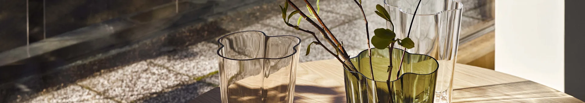 Twee golvende glazen vazen, een heldere en een groene met takken, staan op een lichte houten tafel met schaduwen.