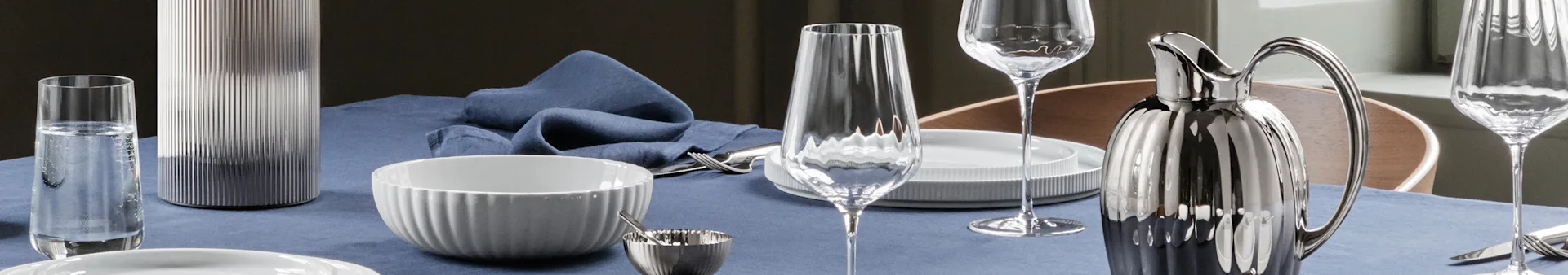 Een eettafel gedekt met een blauw linnen tafelkleed, met witte geribbelde kommen, heldere glazen, zilveren bestek, een zilveren geribbelde kan en een grijze geribbelde vaas.