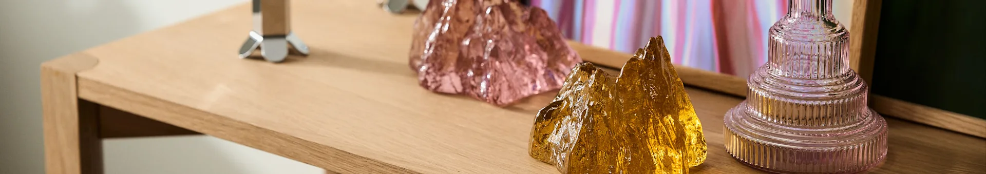 Decoratieve roze en goudgele getextureerde glazen objecten en een geribbelde roze vaas op een lichte houten tafel.