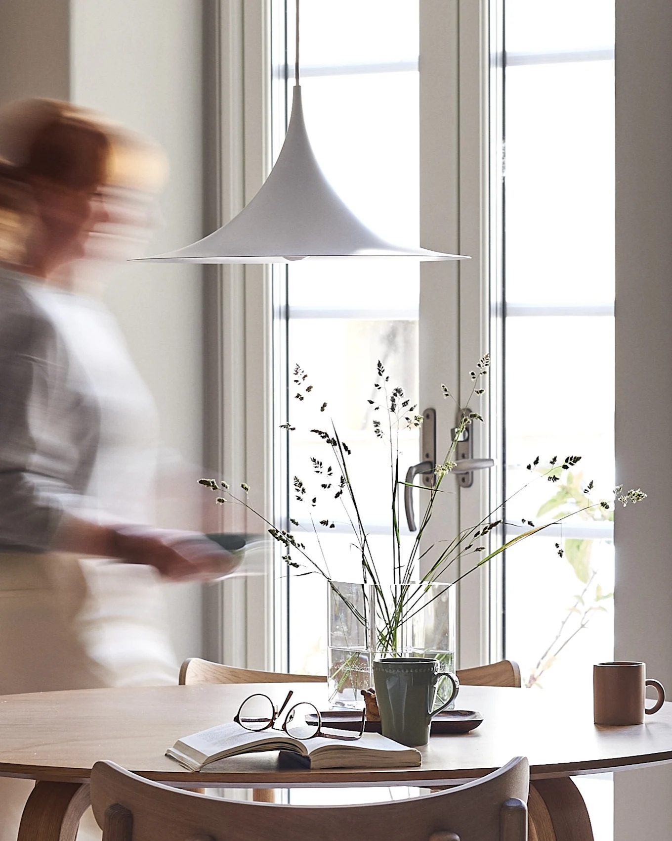 Een witte hanglamp hangt boven een lichte houten tafel met een boek, bril en mokken. Een wazig persoon loopt langs een zonnig raam.