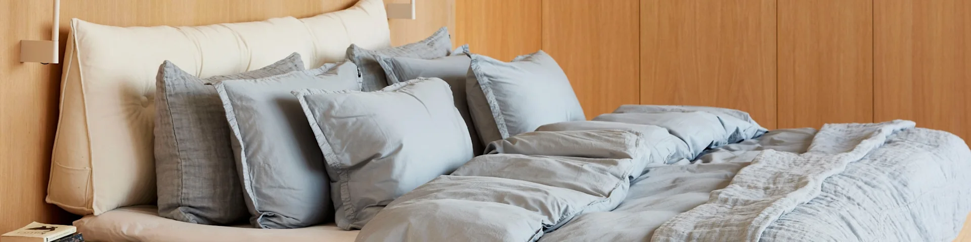Bed met gelaagd beddengoed: een gebroken wit kussen, verschillende lichtgrijze kussens, een grijs dekbed en een getextureerde lichtgrijze plaid. Een licht houten lambrisering bevindt zich erachter.