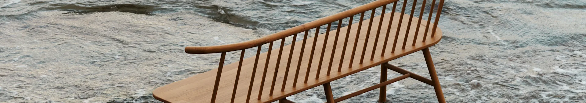 Een lichte houten bank met spijlenrug en gebogen rugleuning, deels ondergedompeld in ondiep, rimpelend water.