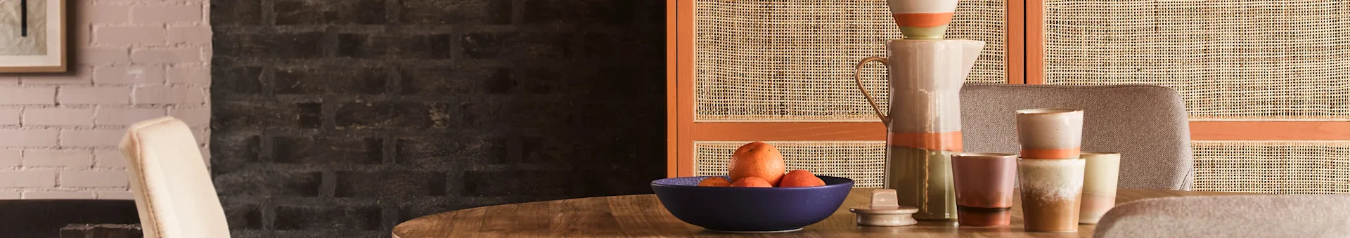 Een eettafel met een blauwe schaal met citrusvruchten en wortelen, omringd door keramische kannen en mokken, tegen een rieten scherm en bakstenen muur.