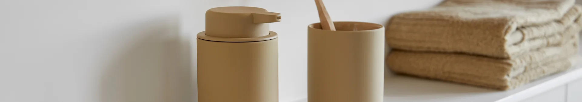 Beige zeepdispenser en tandenborstelhouder met houten tandenborstel, naast gestapelde beige handdoeken op wit oppervlak.
