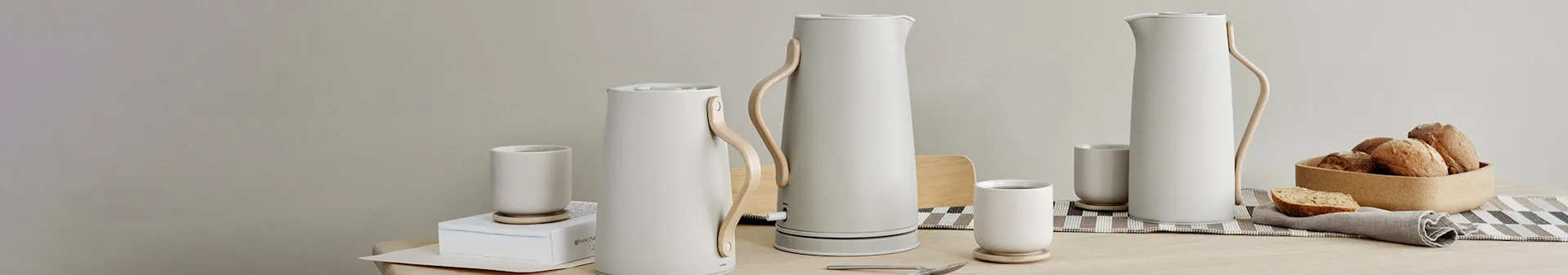 Minimalistische tafelsetting met drie witte kannen, bijpassende mokken en een broodmand op een houten tafel.