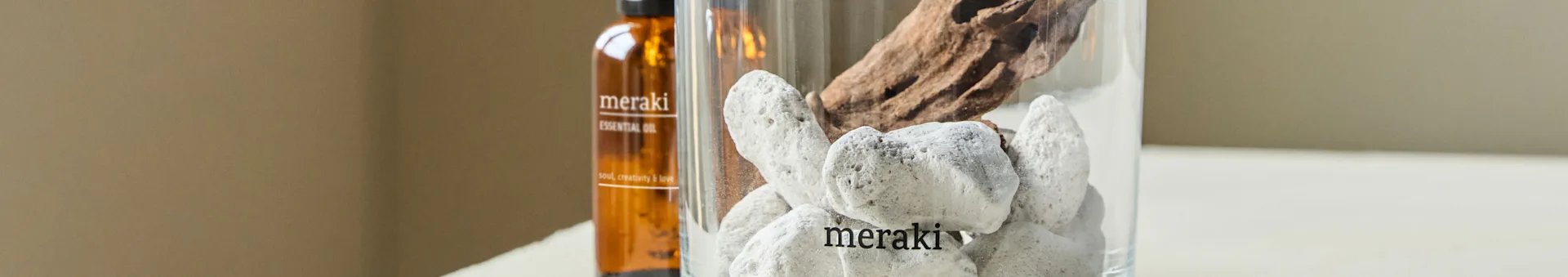 Een bruine etherische oliefles en een heldere vaas met witte stenen en drijfhout. Zowel de fles als de vaas zijn gelabeld met 'meraki'.