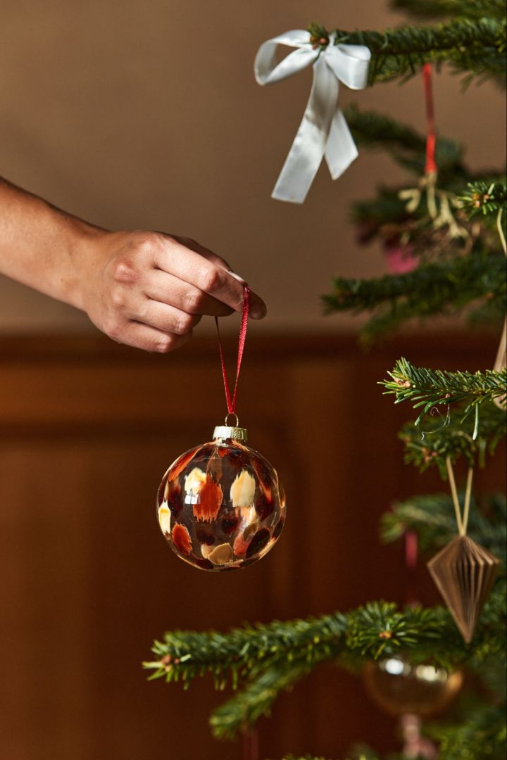 Een hand houdt een glazen kerstbal in de boom. Glazen decoraties blijven voor Kerstmis 2024 even populair als altijd.