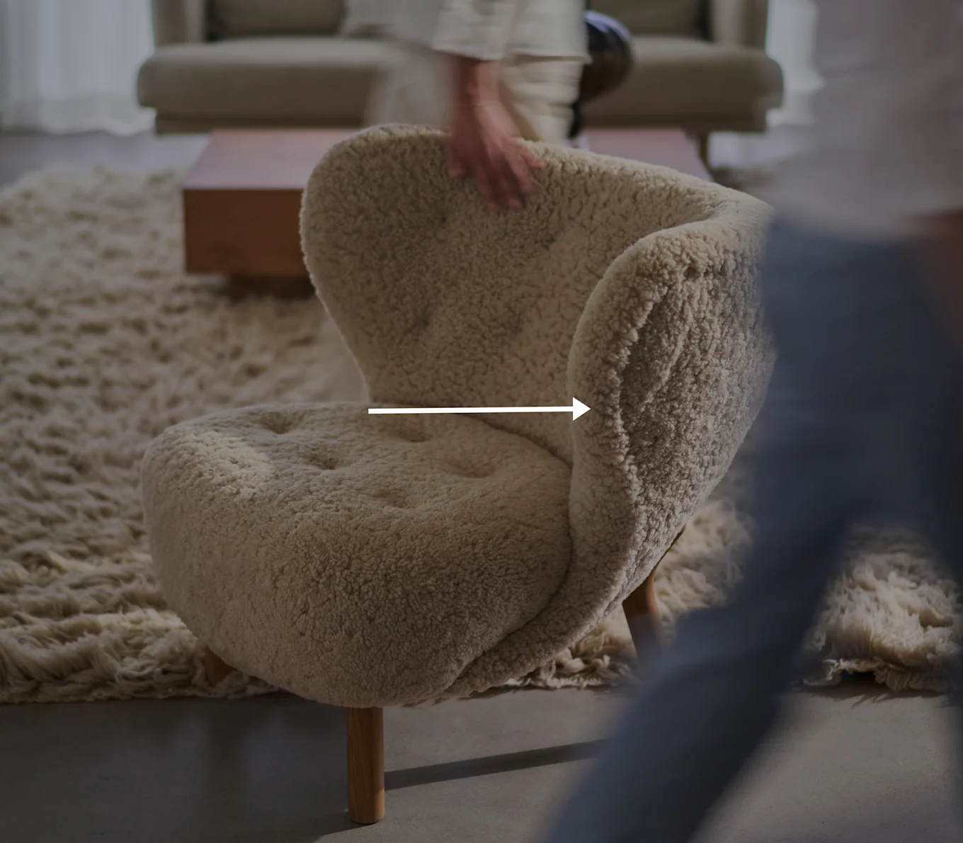 Crèmekleurige, pluizige fauteuil met houten poten in een woonkamer, een vage hand raakt de rugleuning aan.