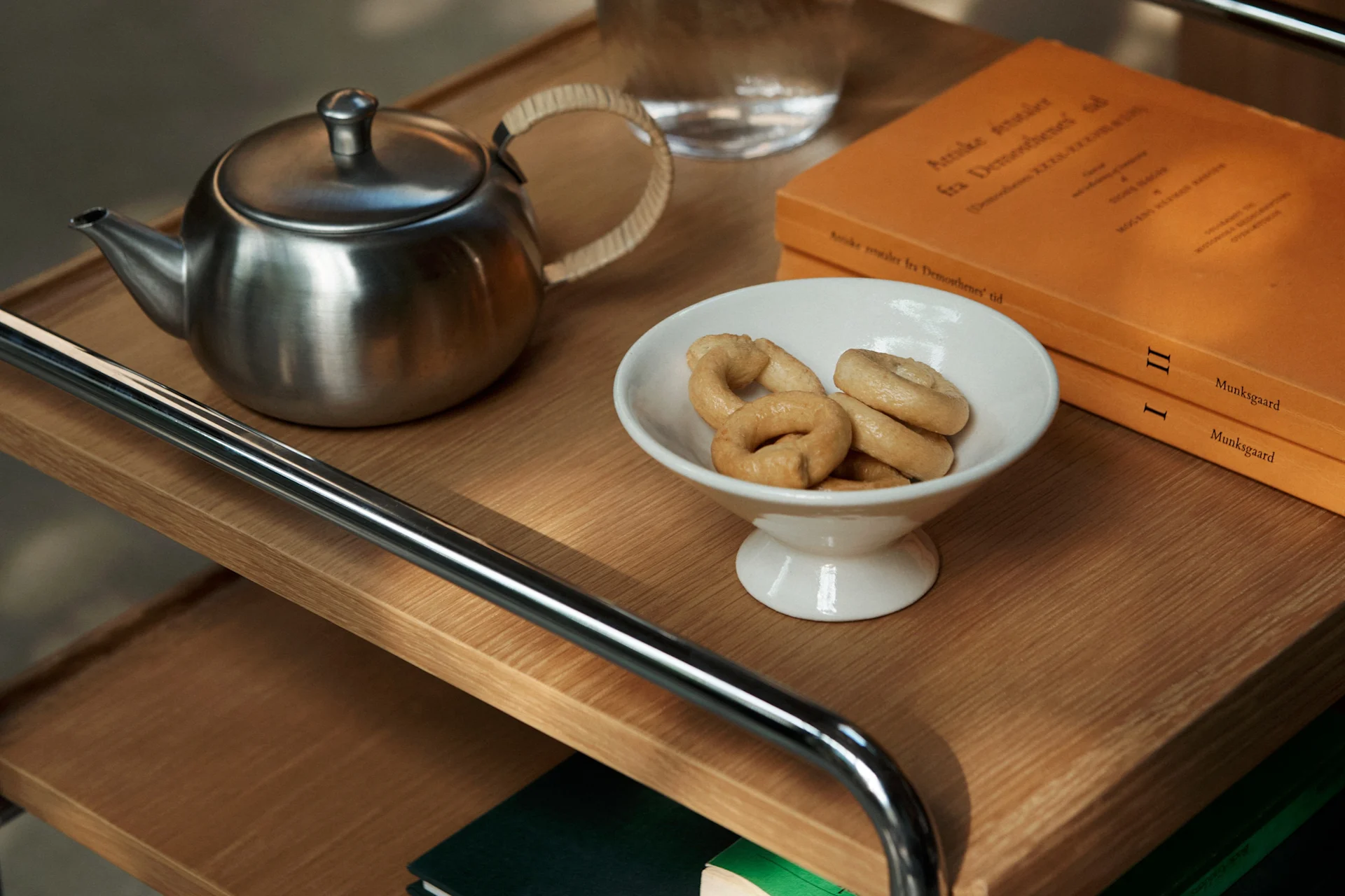 Een roestvrijstalen theepot, een witte kom met ringkoekjes en twee oranje boeken op een houten plank.