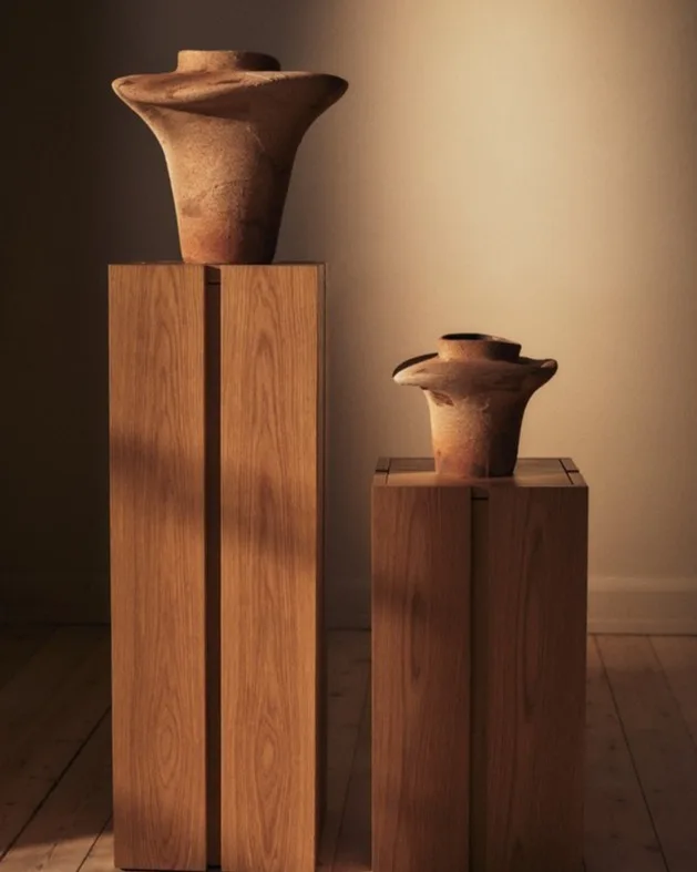 Twee aardekleurige, uitlopende keramische vazen op lichte houten sokkels.