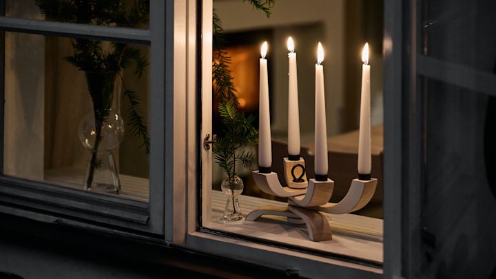 In de vensterbank staat de elegante Nordic Light kandelaar van Design House Stockholm. Op de achtergrond is een interieur zichtbaar met een knusse open haard en kerstgroen in een vaas.