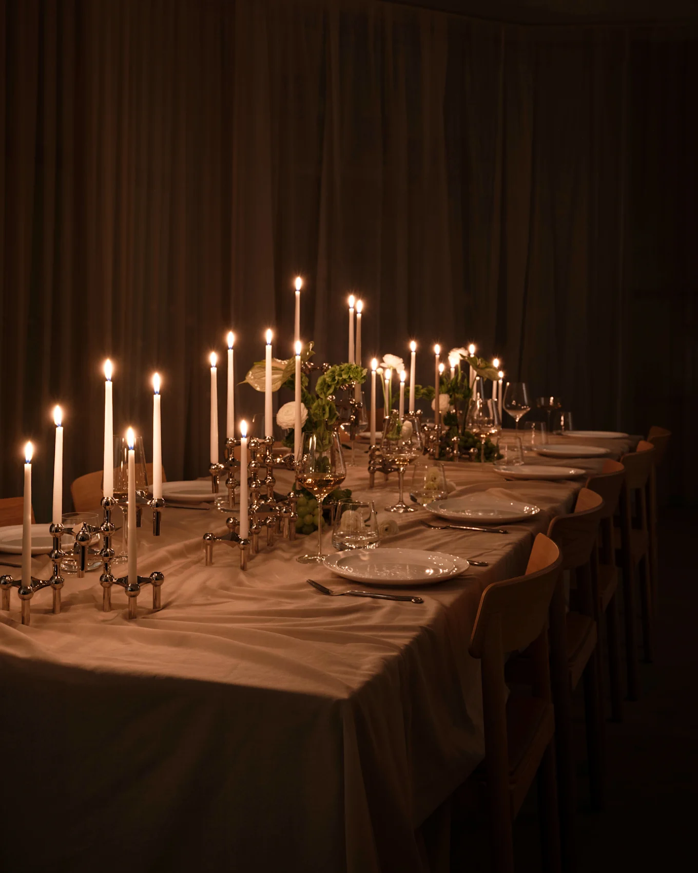 Lange eettafel met brandende kaarsen in zilveren houders, witte borden en glazen, warme gloed in schemerig verlichte kamer.