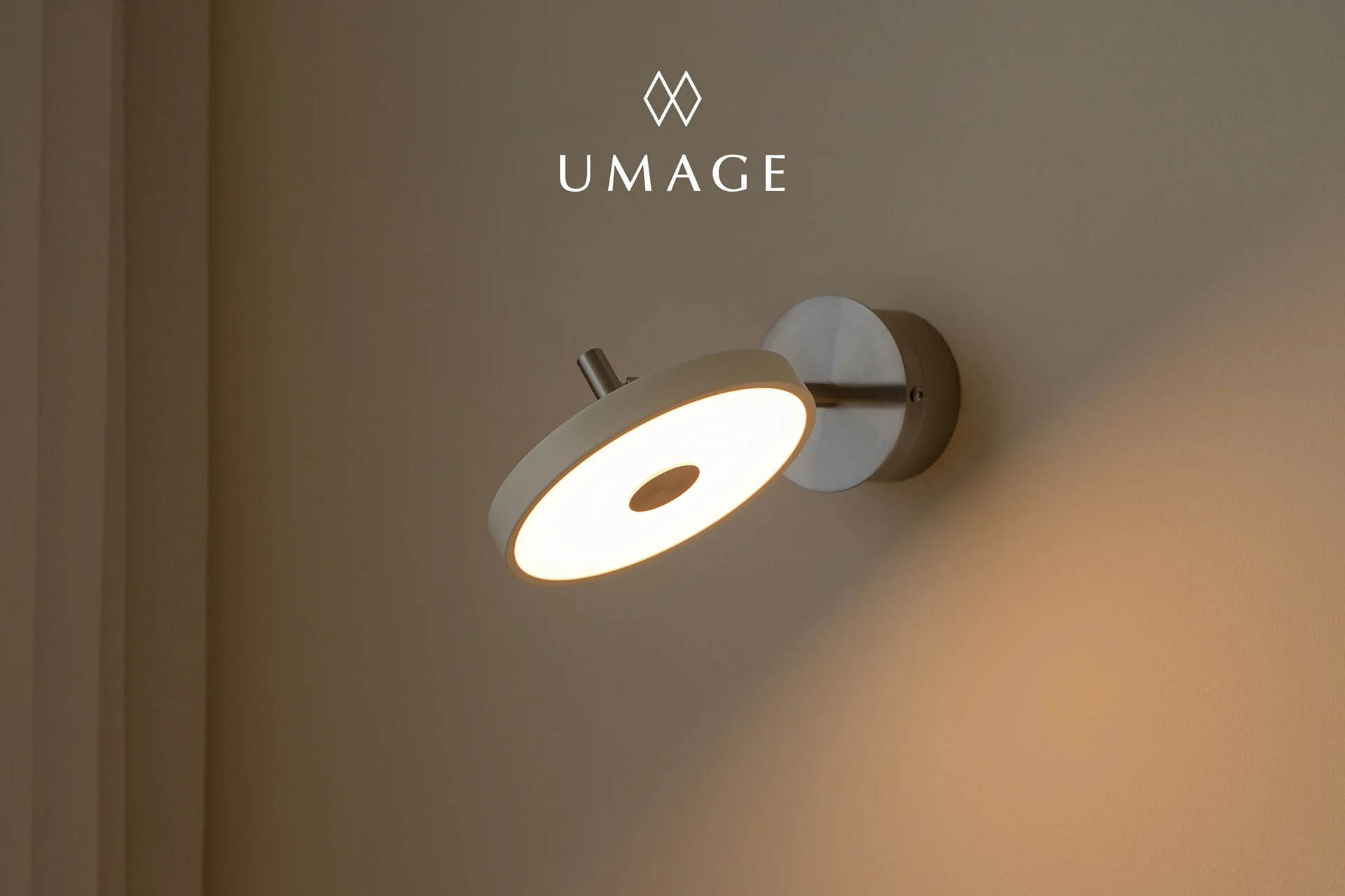 Een moderne ronde wandlamp met warm gloeiend licht, gemonteerd op een lichtbruine muur. UMA-logo erboven.