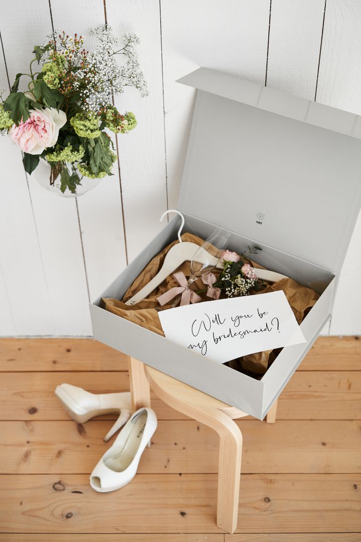 Verras je maid of honour met een cadeaubox. Hier zie je een cadeaubox op een kruk, gevuld met een gepersonaliseerde kledinghanger, champagneglas en een bosje bloemen. Op de grond liggen een paar witte hakken en er hangt een bos bloemen aan de muur.