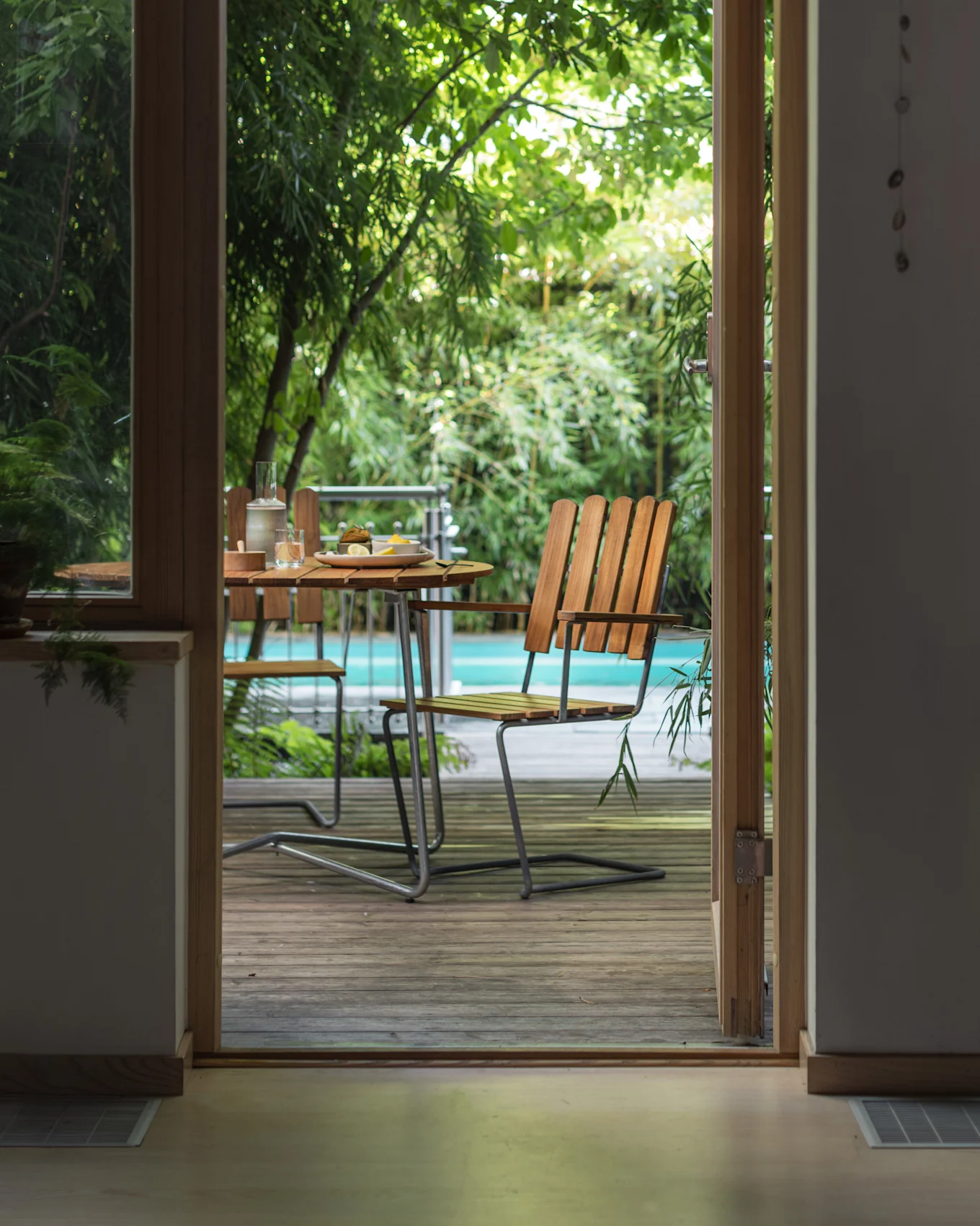 Uitzicht door een open deur naar een buitenpatio met een houten tafel, stoelen en een zwembad omringd door groene planten.