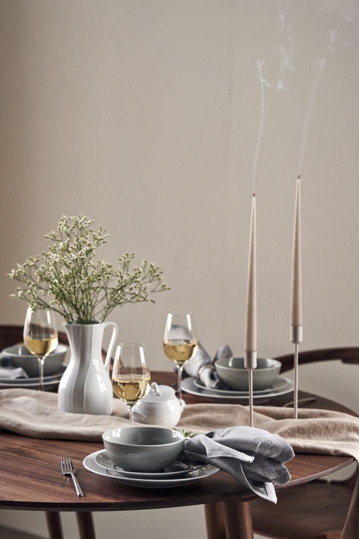 Een feestelijk gedekte tafel met kaarsen, weidebloemen in een karaf van Pli Blanc en Zweeds Grace porselein in wit en grijs. 