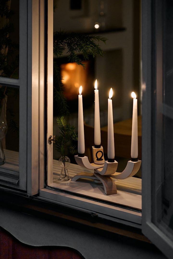 In de vensterbank staat de elegante Nordic Light kandelaar van Design House Stockholm. Op de achtergrond is een interieur zichtbaar met een knusse open haard en kerstgroen in een vaas.