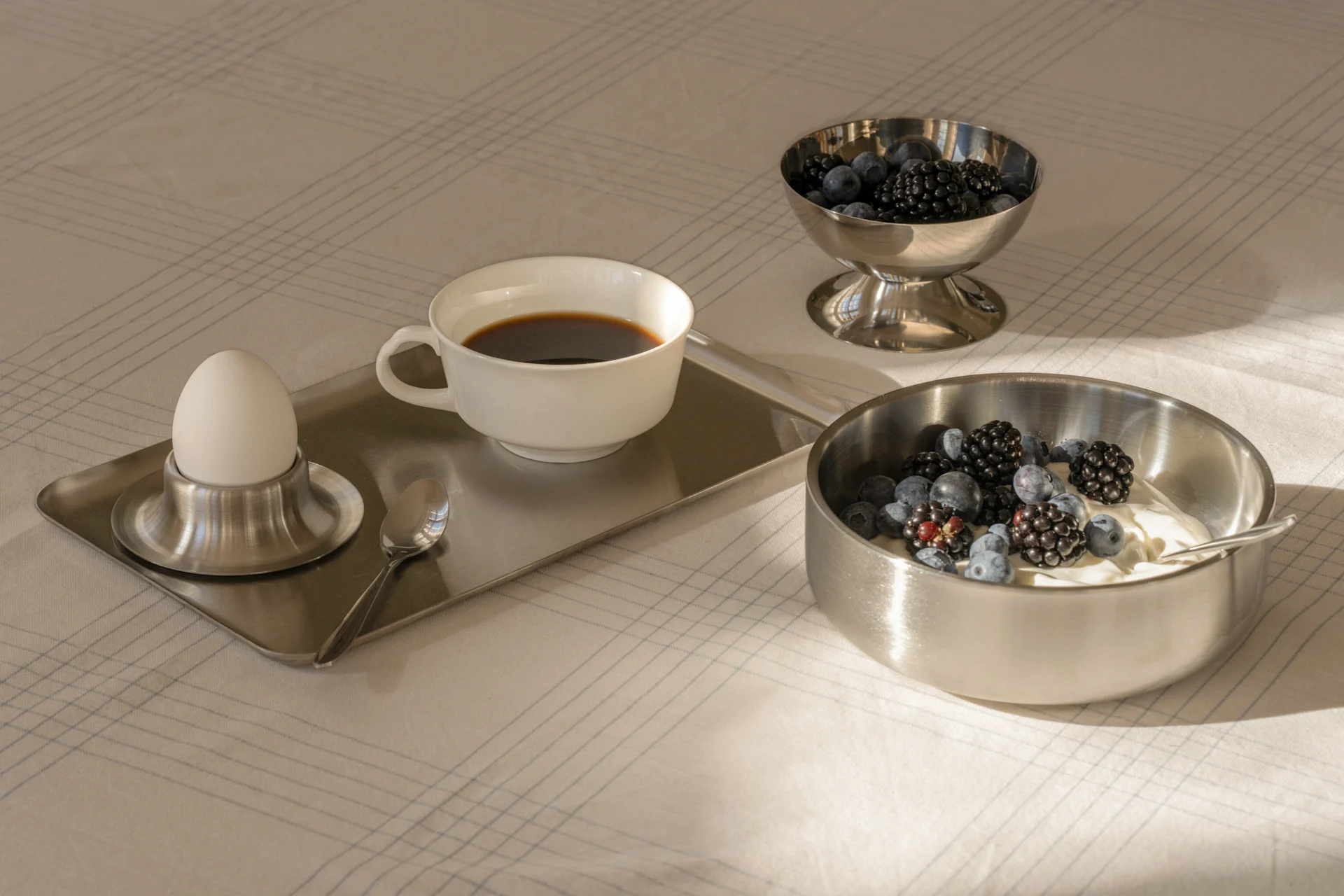 Ontbijt met koffie, gekookt ei, yoghurt en verse bessen in zilveren schalen op een gedessineerd tafelkleed.