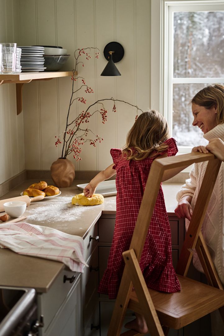 Men ziet een  warme, huiselijke sfeer  waarin een meisje op een stoel deeg uitrolt, terwijl haar moeder glimlachend toekijkt in een sfeervolle keuken met winterse decoraties en een besneeuwd uitzicht.