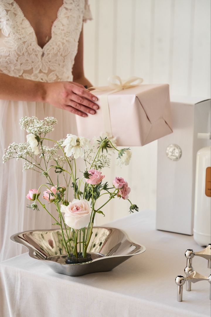 Elke bruiloft heeft een plek nodig waar de gasten hun cadeau kunnen neerleggen. Hier zie je een deel van een tafel met een bloemenarrangement in de Alvar Aalto schaal. Op de achtergrond zie je de bruid die een ingepakt cadeau vasthoudt.