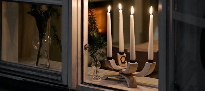 In de vensterbank staat de elegante Nordic Light kandelaar van Design House Stockholm. Op de achtergrond is een interieur zichtbaar met een knusse open haard en kerstgroen in een vaas.