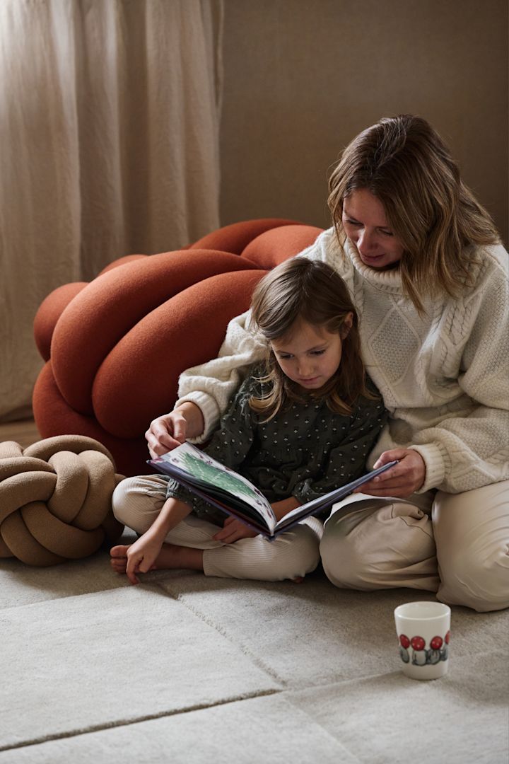 Een moeder en dochter zitten op de grond, verdiept in een boek, omringd door kleurrijke Knot kussens van Design House Stockholm. Naast hen staat een Children of the Forest mok.