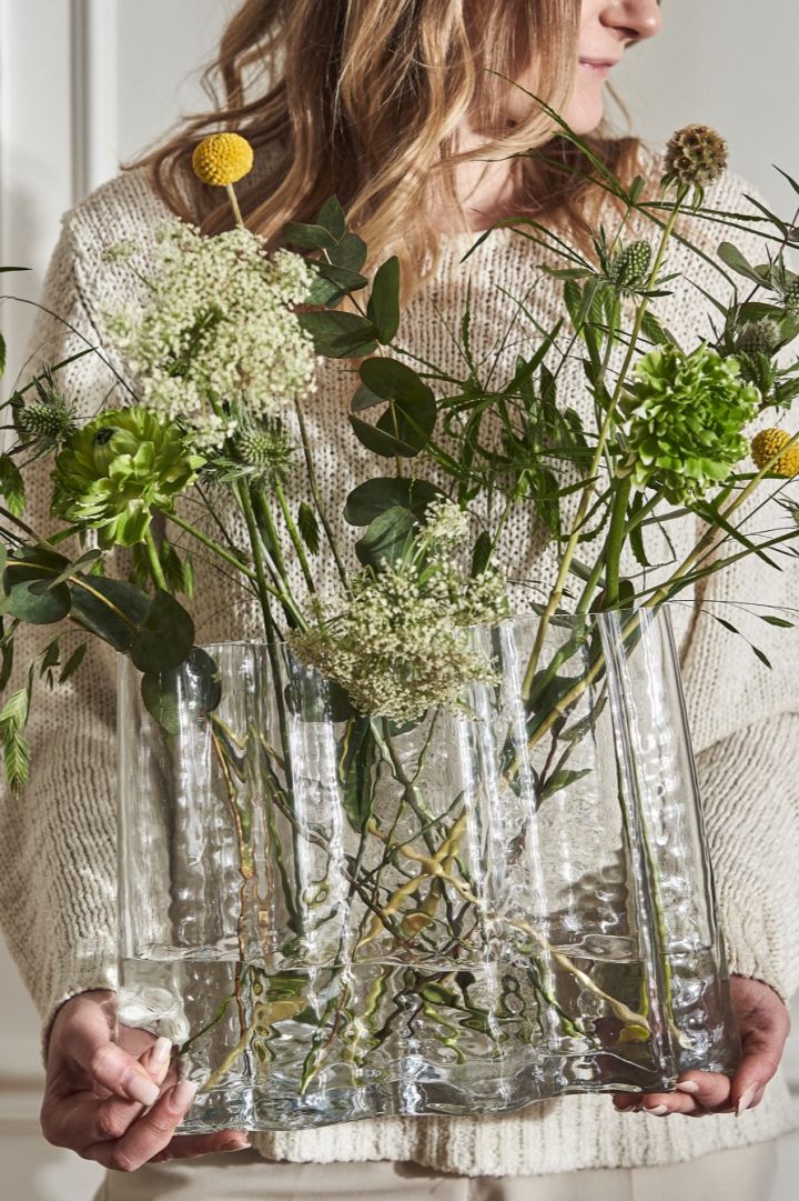 Een vrouw gekleed in lichte kleuren houdt de Gry-vaas vast, die gevuld is met wilde lentebloemen. 