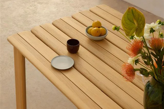 Licht houten latten tafel met een kom citroenen, donkere mok, klein bord en een vaas met oranje en witte bloemen.