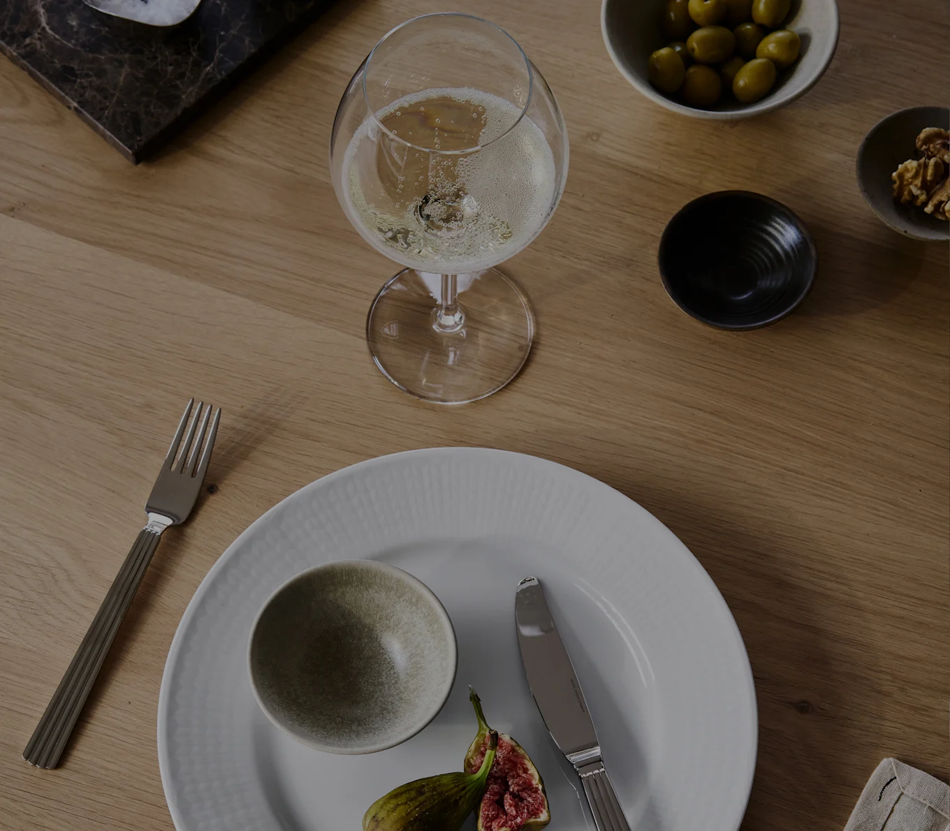 Een glas champagne, wit bord met een kom en vijgen, vork, mes, olijven en walnoten op een houten tafel.