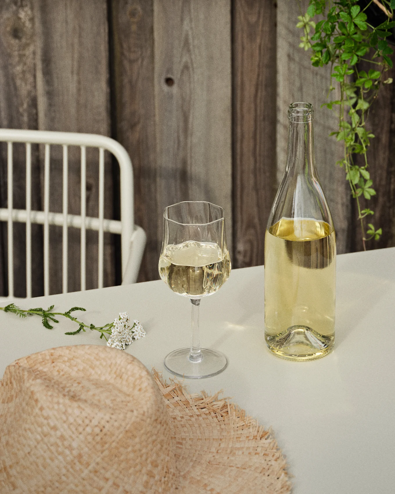 Een fles en een glas witte wijn op een tafel met een strohoed en witte bloemen, tegen een houten muur en groene klimplanten.