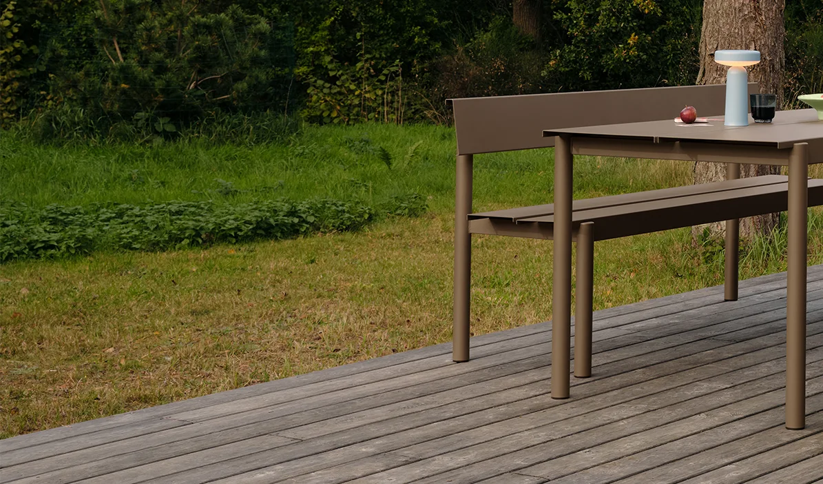 Moderne taupekleurige buiten eettafel en bank op een houten vlonder, met een draagbare lamp en appels op tafel.