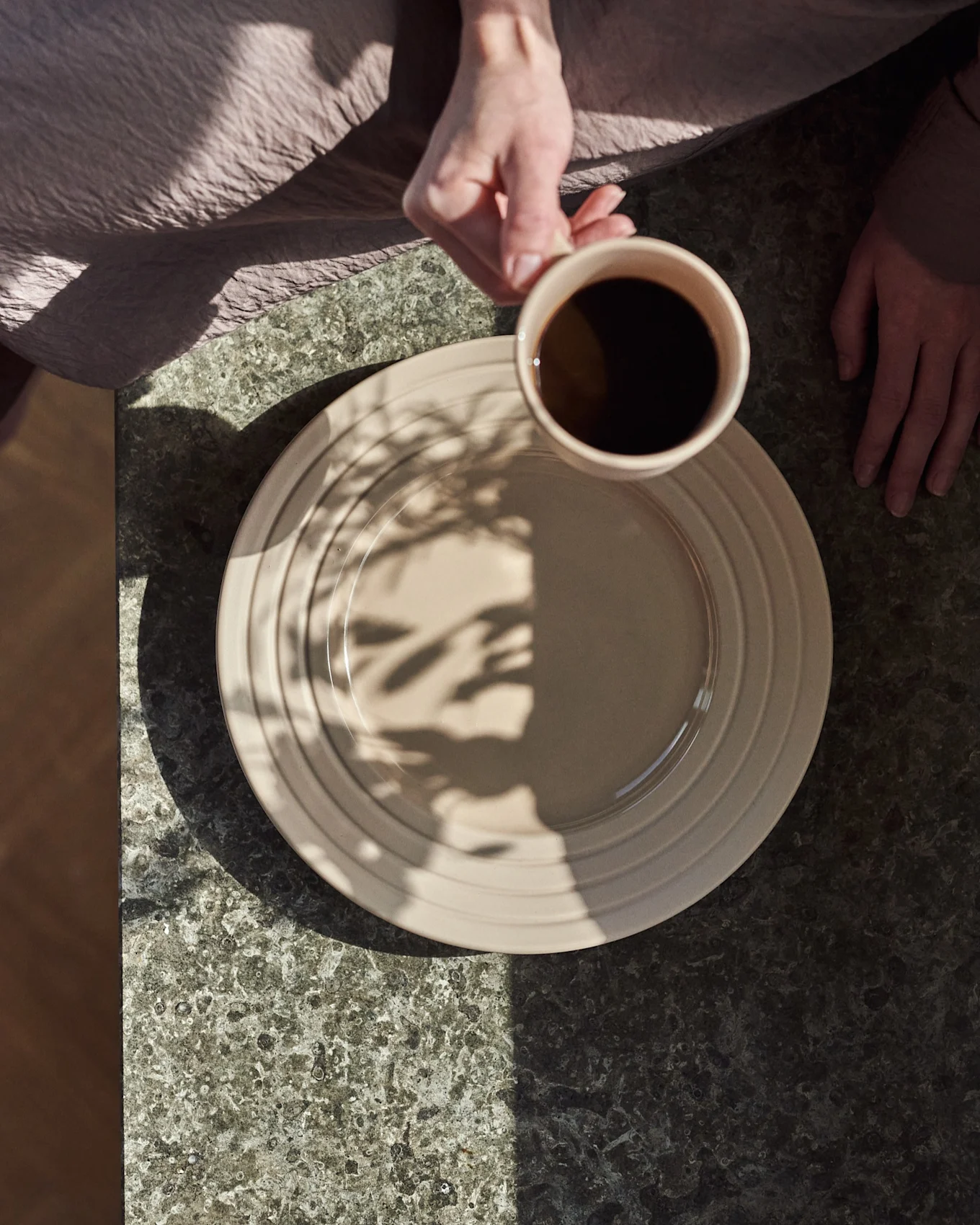 Bovenaanzicht: handen, één houdt een beige mok met zwarte koffie boven een beige bord op een donkere stenen tafel, met bladschaduwen.