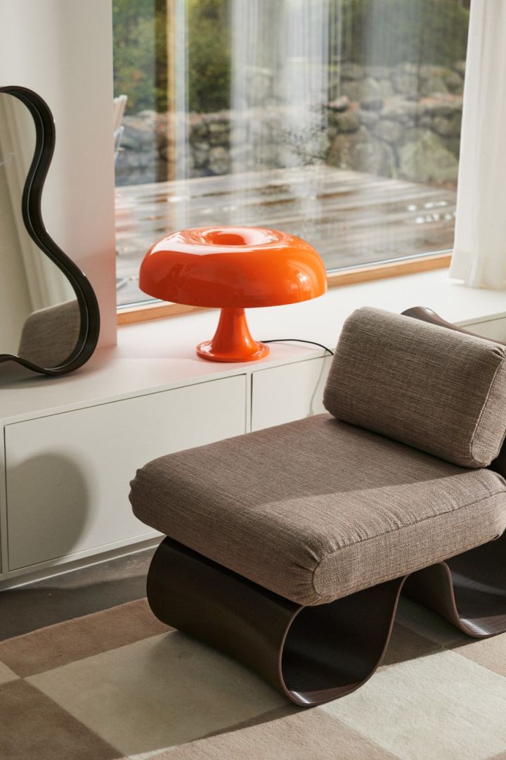 De oranje Nessino-lamp met een donutvormige kap staat in een raam, naast een spiegel met gebogen randen en een bijpassende fauteuil.