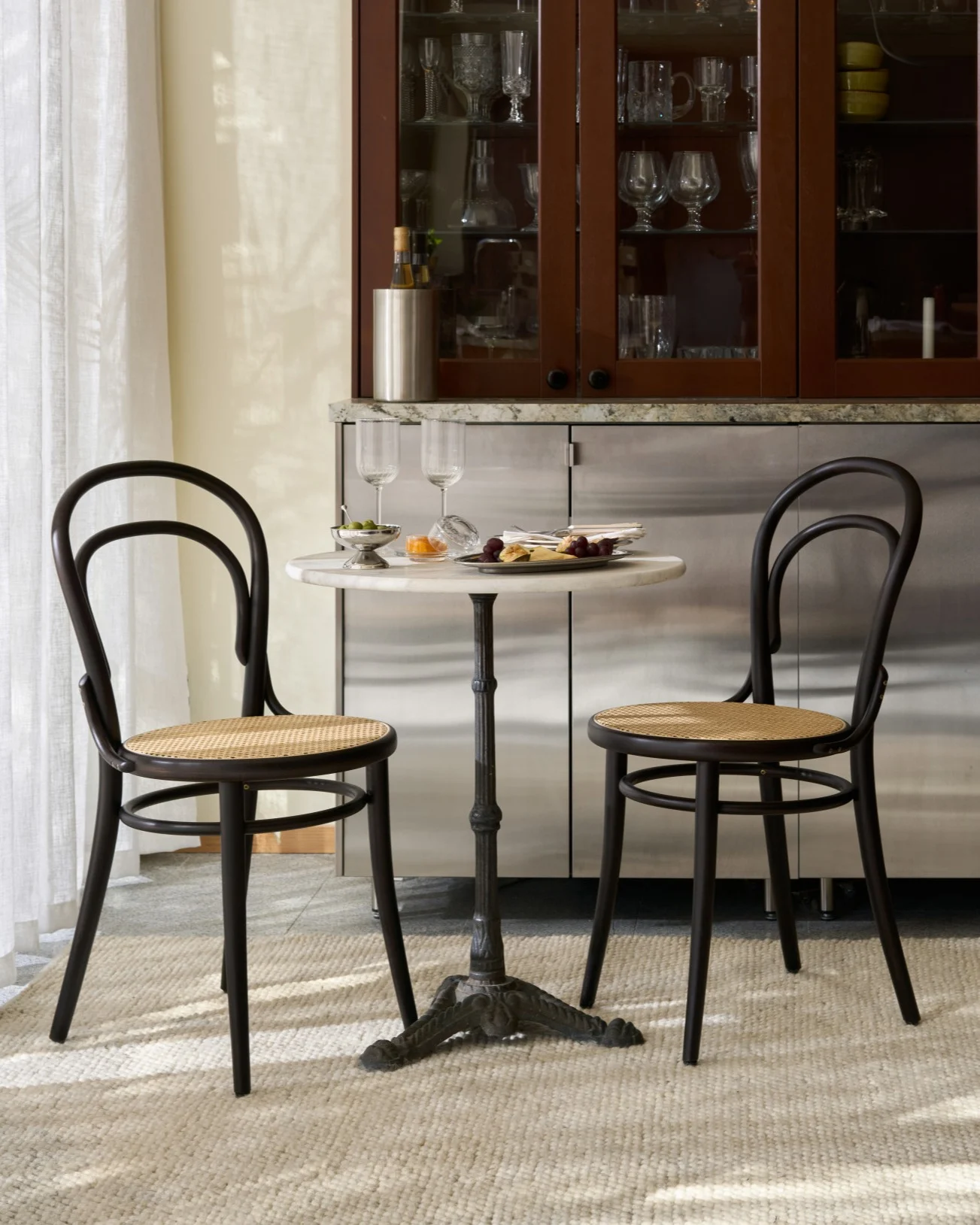 Een bistrotafel met twee donkere houten en rieten stoelen, met hapjes en wijnglazen, voor een glazen kast.