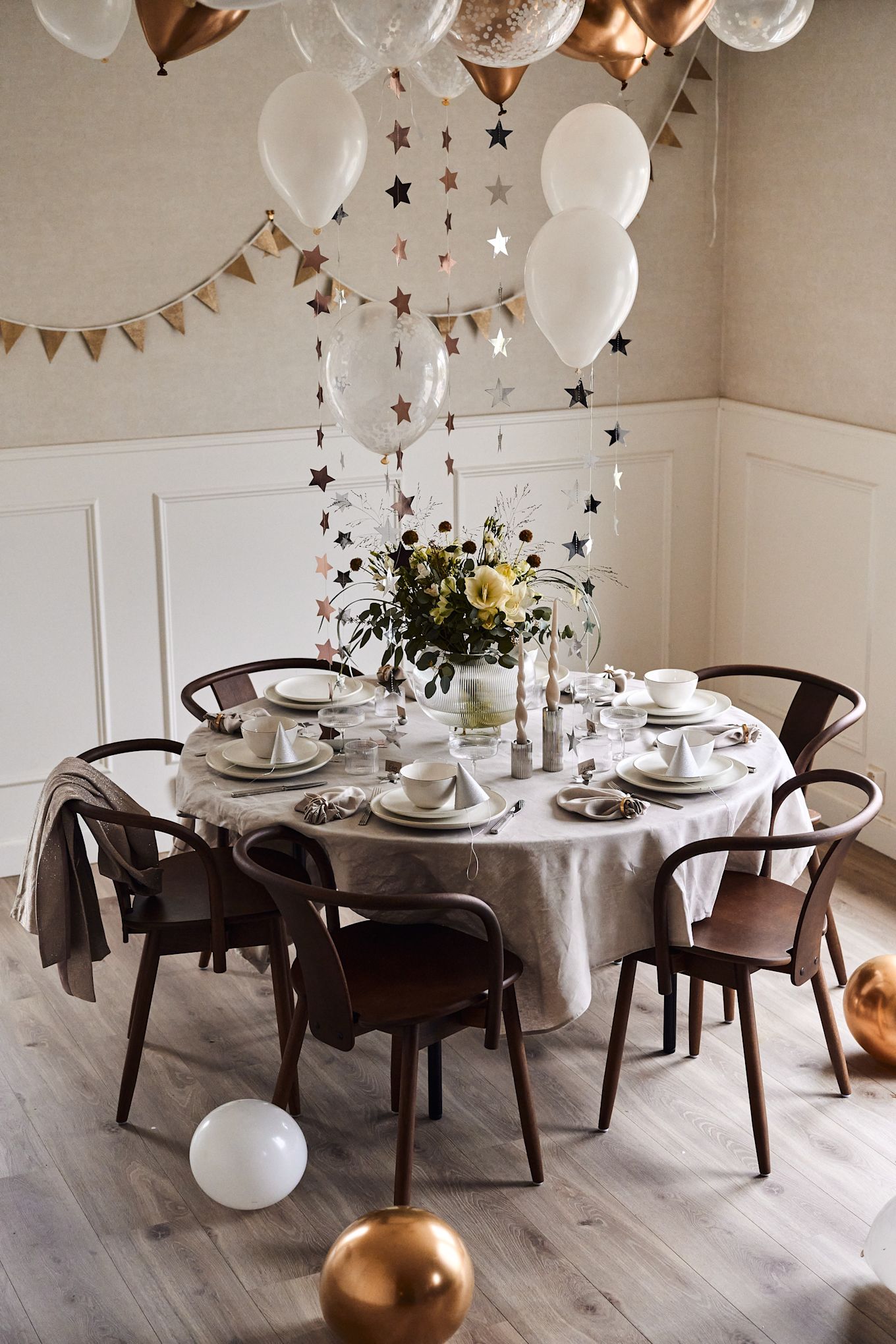 Ideeën voor je nieuwjaarsfeest: Versier de tafel met ballonnen en sterren in wit en goud die aan het plafond hangen.