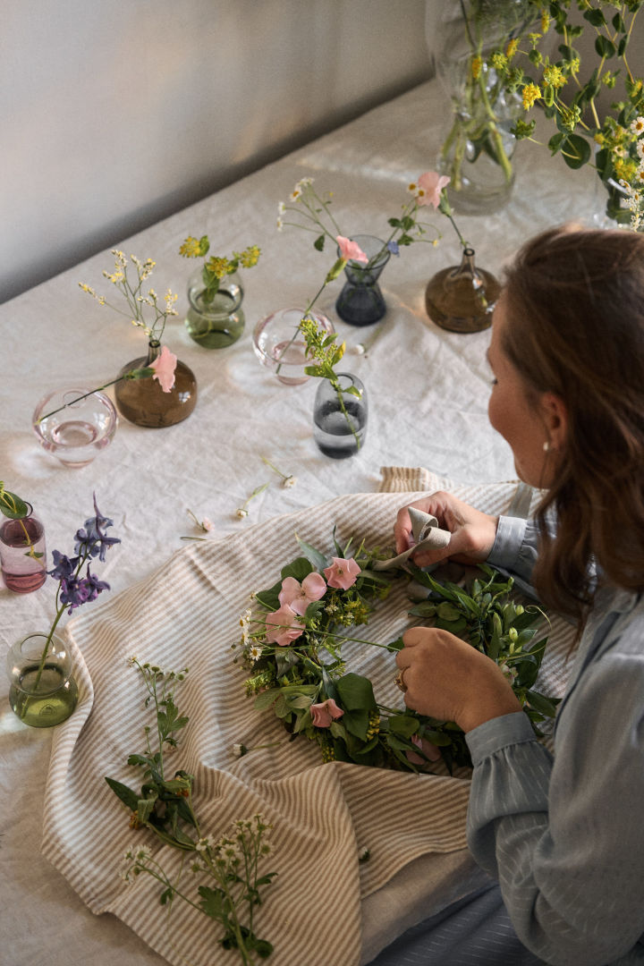 Een Midzomertraditie in Zweden is het maken van een bloemenkrans van bloemen die je zelf hebt geplukt. Hier zie je een vrouw aan een tafel zitten terwijl ze een bloemenkrans maakt, omringd door talloze mini Midzomer vaasjes van Orrefors.