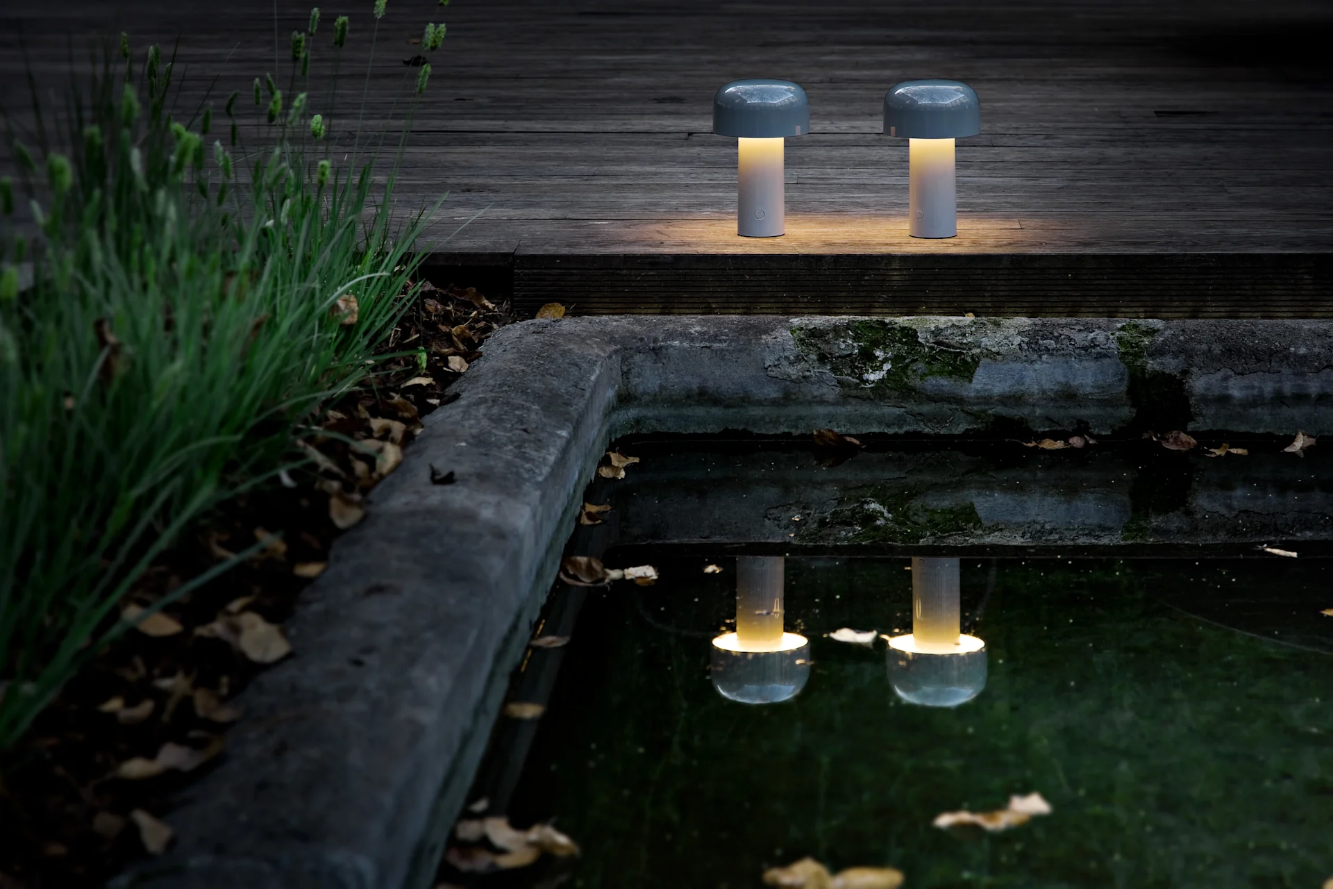 Twee draagbare paddestoelvormige tafellampen met warm geel licht staan op een donker houten dek bij een vijver, hun gloed reflecterend.