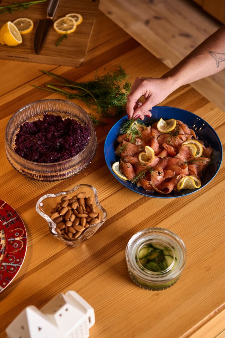 Zweeds kerstdiner: Een hand legt een stuk dille op een blauw bord met zalm en citroen, een traditioneel gerecht op een Zweeds kerstfeest. Ernaast staan schaaltjes met rode kool en amandelen.