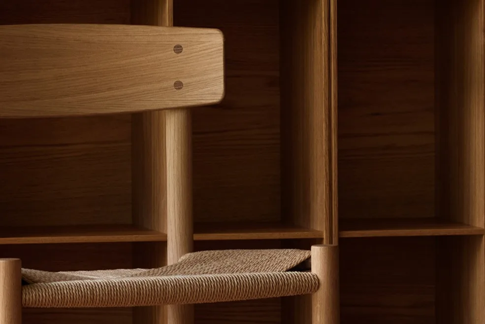 Een lichte houten stoel met een gevlochten zitting voor een wand met vierkante houten planken en een patroonhouten vloer.