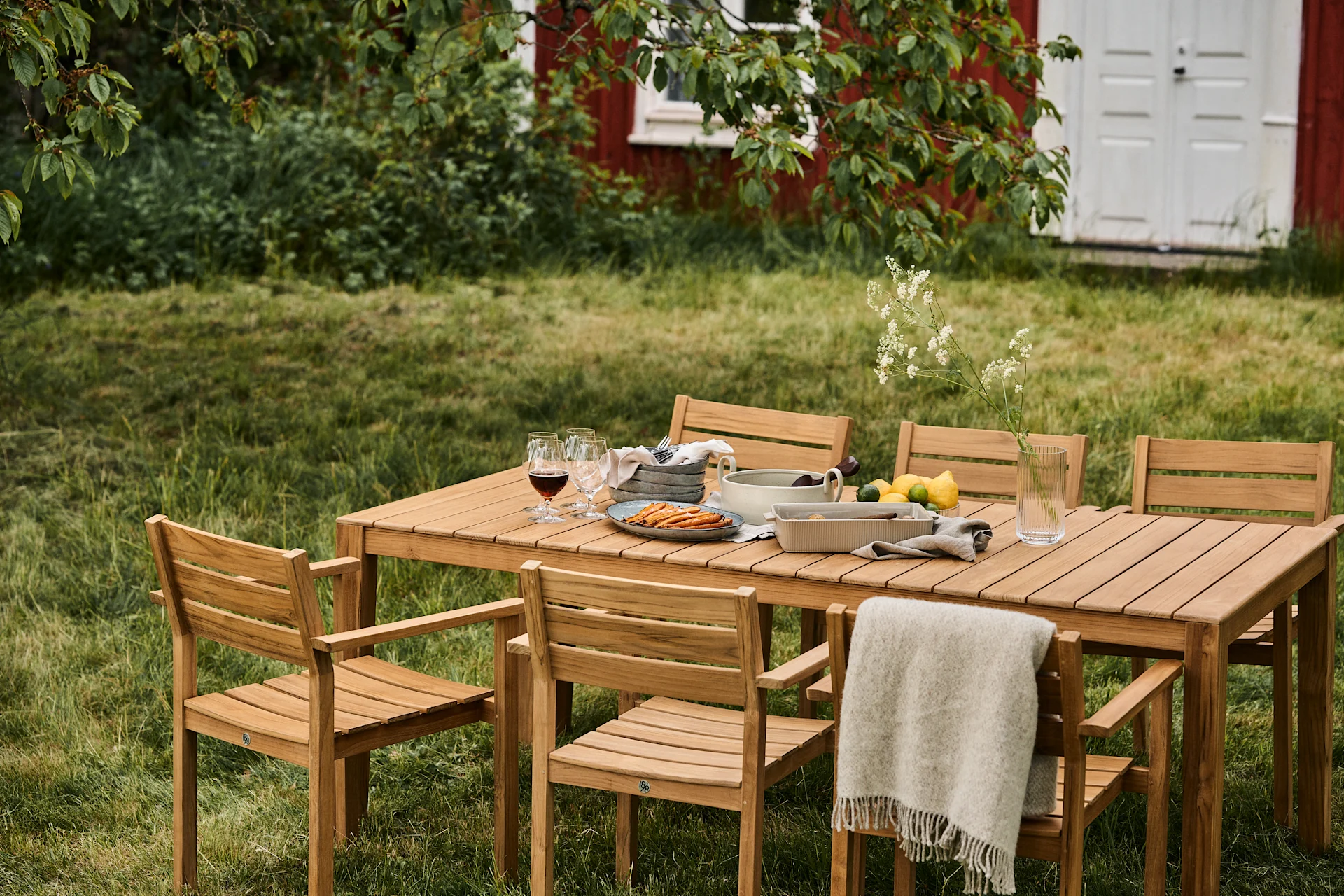 Buiten eethoek met houten tafel en zes stoelen op een gazon, gedekt met eten, drinken en een plaid, met een rood huis op de achtergrond.