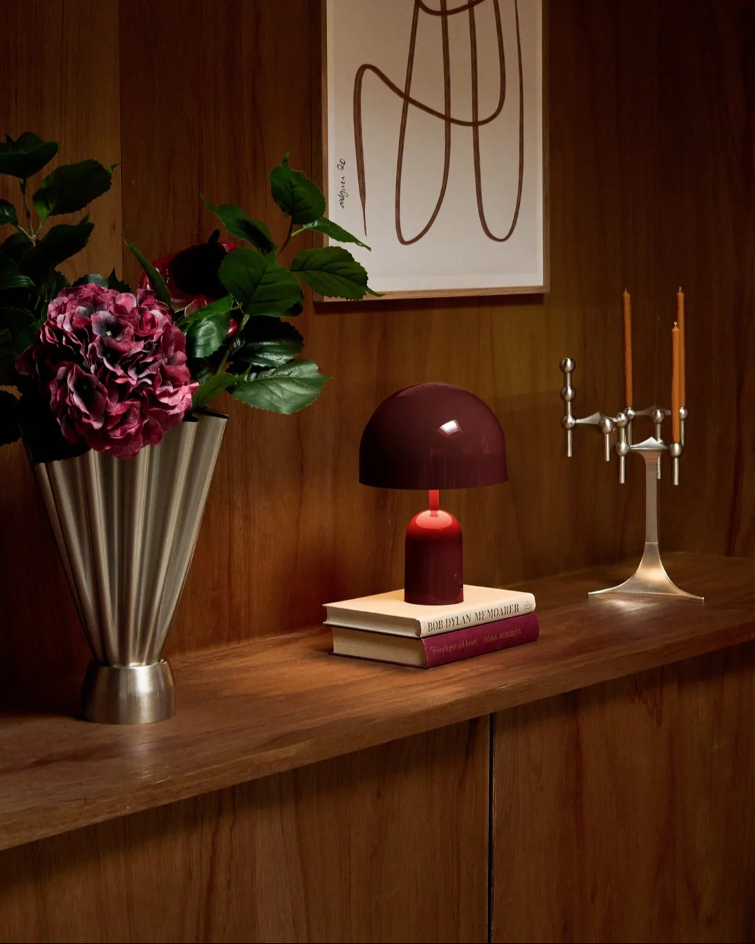 Gezellige houten plank met zilveren geribbelde vaas, dieprode hortensia's, donkerrode lamp op boeken en zilveren kandelaar.