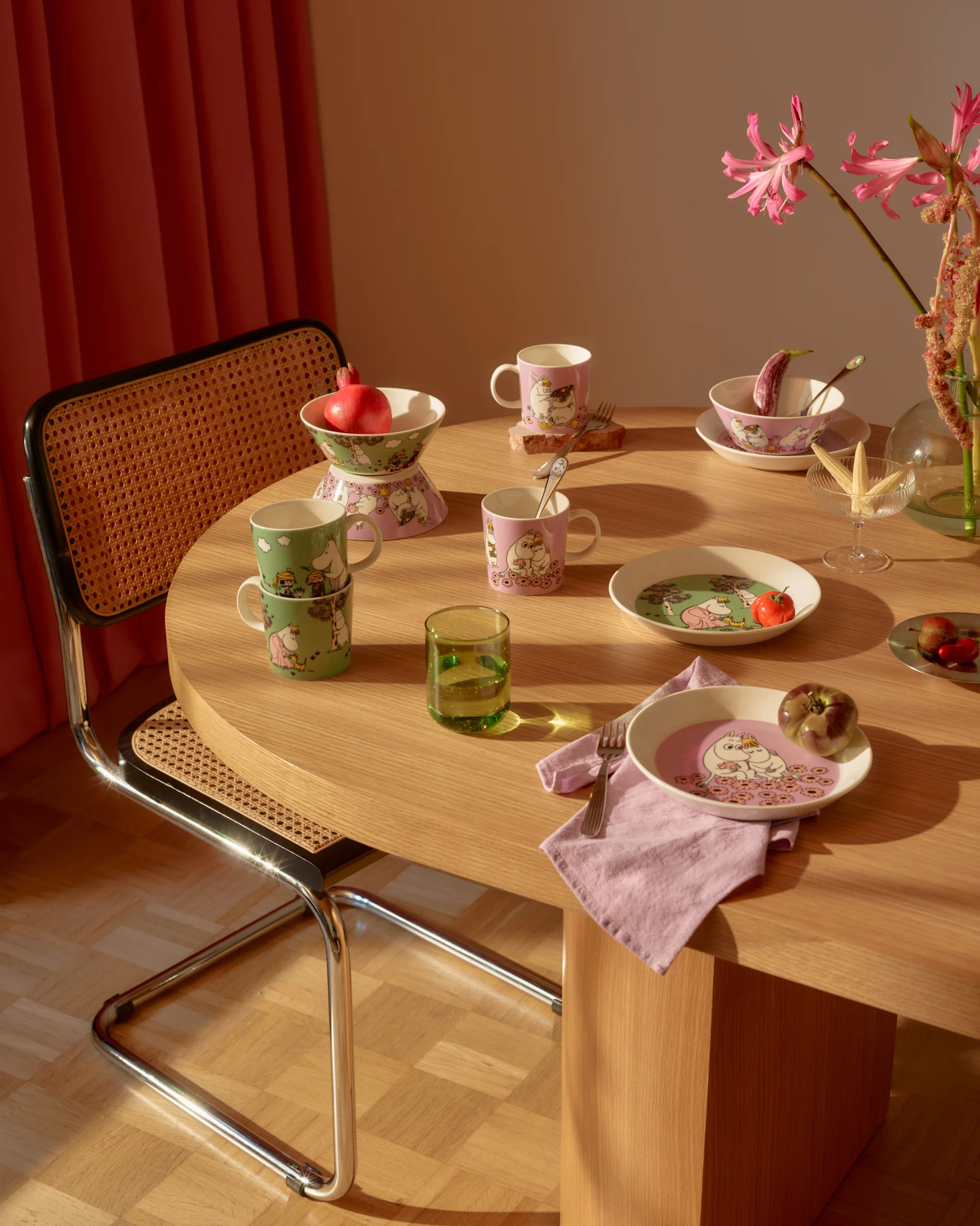 Eettafel met roze en groen Moomin-servies, vers fruit, een groen glas en roze bloemen.
