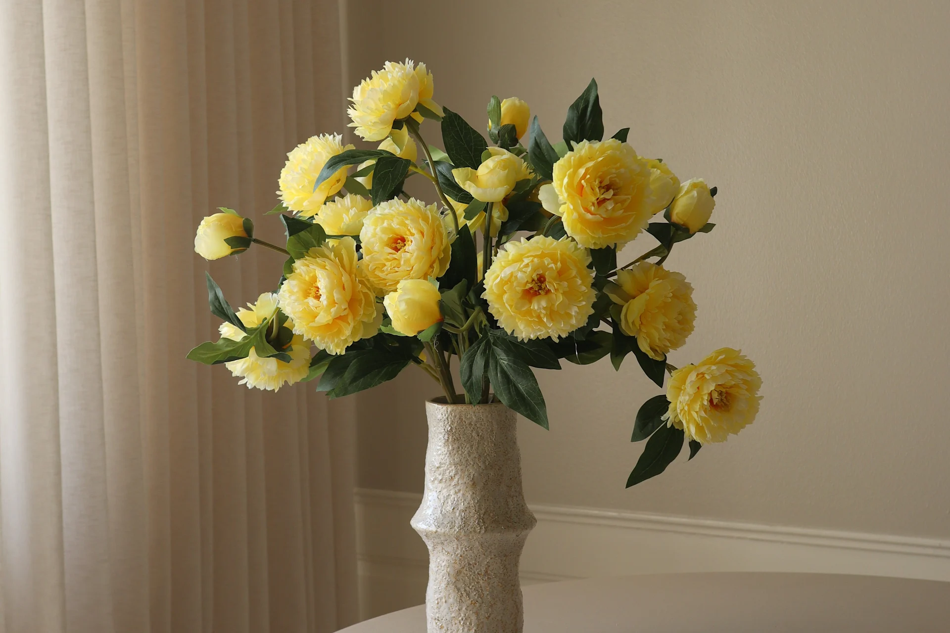 Een lichtgekleurde, gestructureerde vaas bevat een levendig boeket van gele pioenachtige bloemen en groene bladeren tegen een lichte achtergrond.