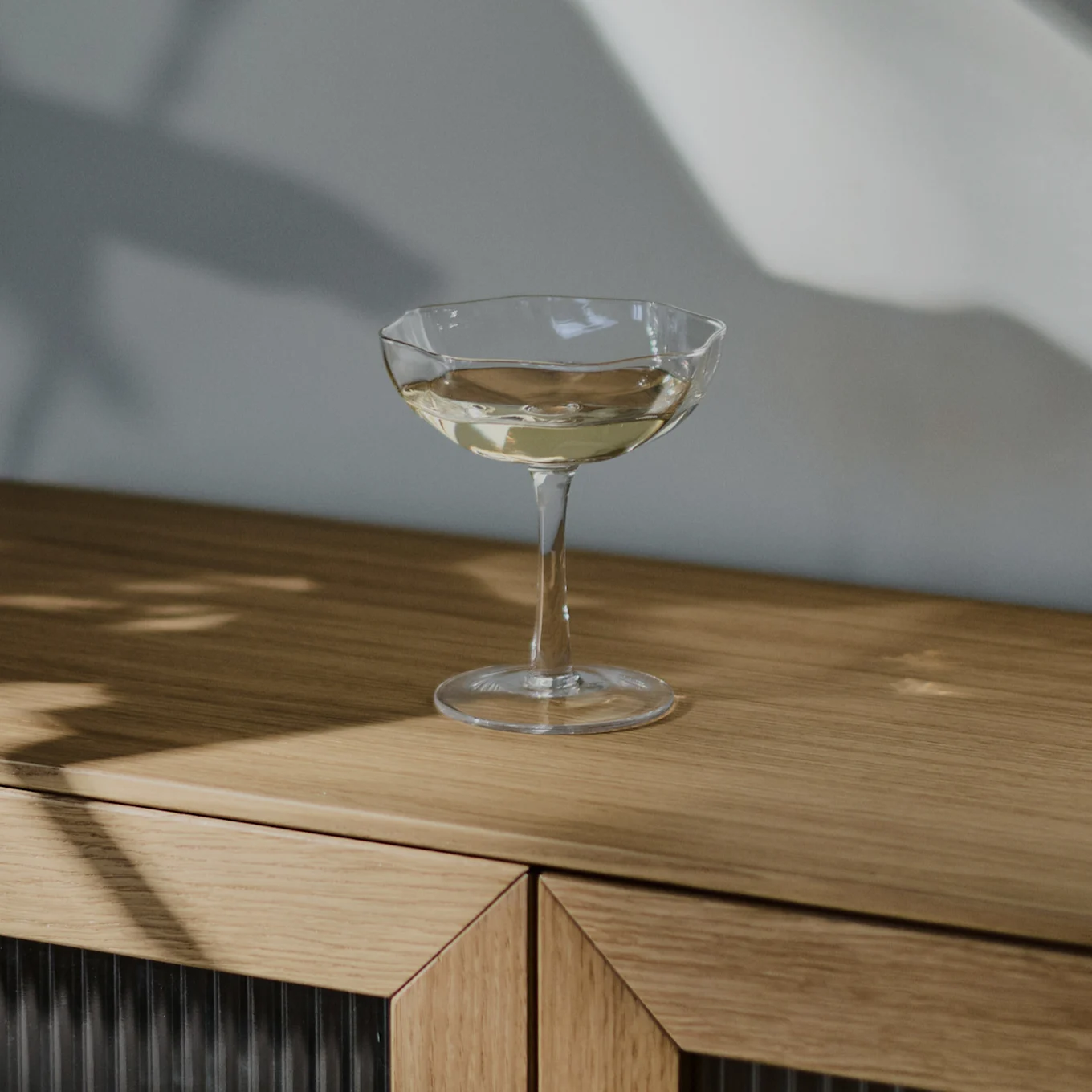 Een helder coupeglas gevuld met een lichtgekleurde drank staat op een lichtbruin houten dressoir, met schaduwen op de muur.
