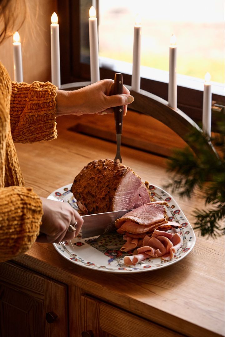 Zweedse kerst: Een vrouw snijdt een traditionele Scandinavische kerstham dicht bij een raam naast een adventskandelaar. 